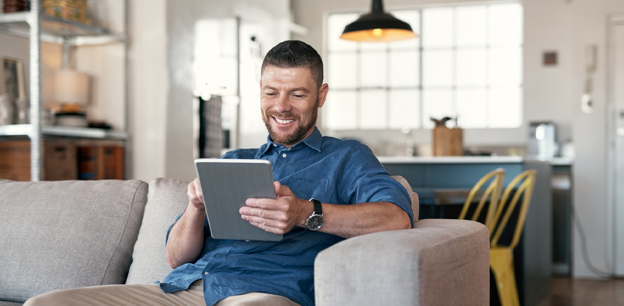 Man smiling while using a tablet on a sofa in a bright living room.
